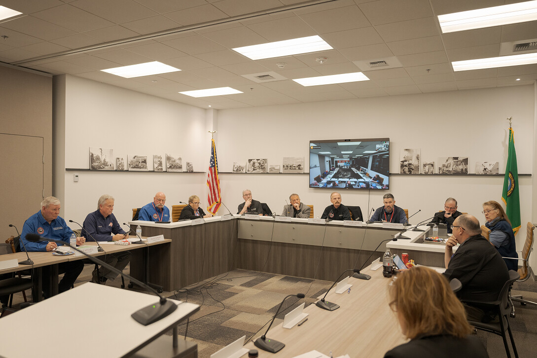 A group of people sit around a U-shaped conference table in a meeting room, with an American flag and a screen displaying a video call in the background.