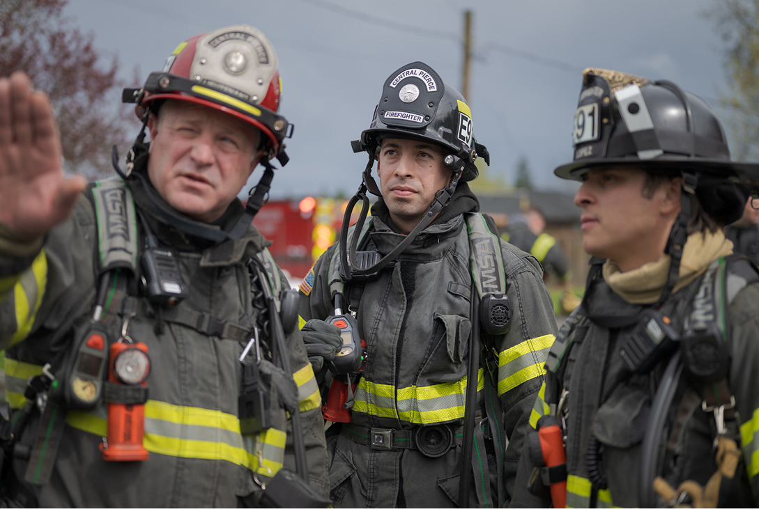 Three firefighters in full gear stand together outdoors; one gestures while the others listen attentively.