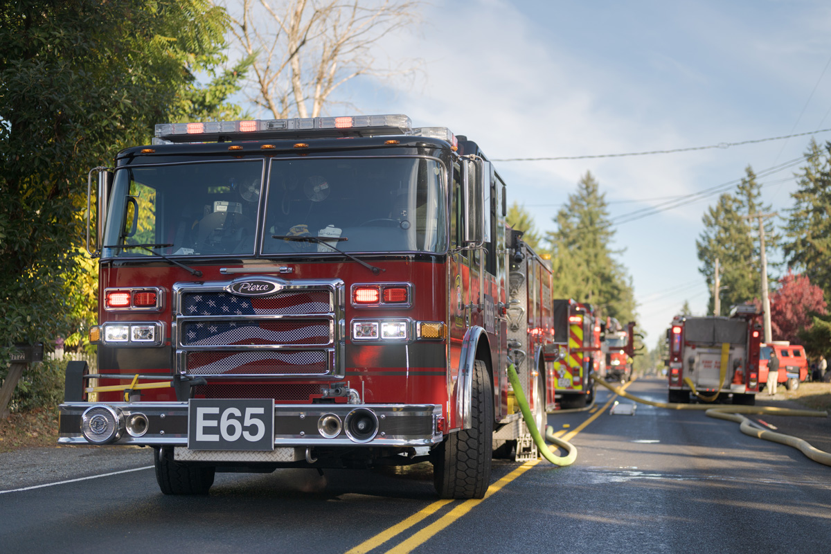 A red fire truck with an American flag design on the grill is parked on a residential street, hoses extended, while additional trucks from Central Pierce Fire & Rescue handle patient care records requests in the background.