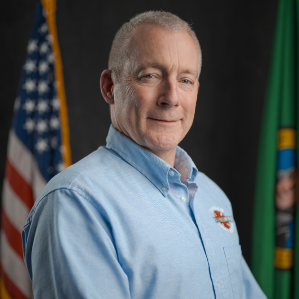 A man in a light blue button-up shirt stands in front of the U.S. flag and another green flag, posing for a formal portrait.