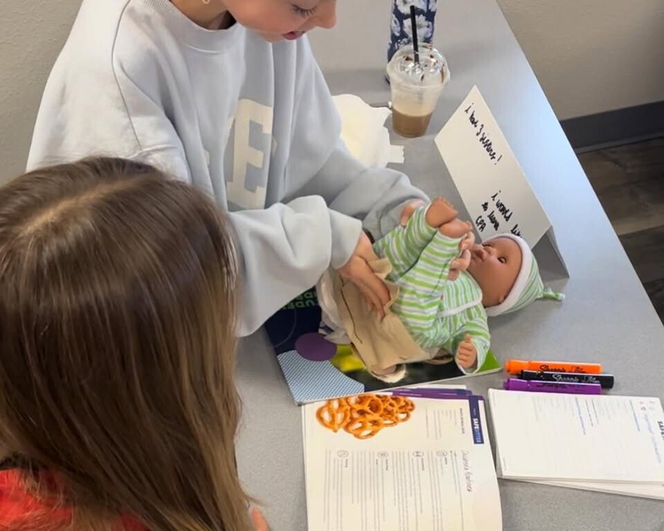 Two young people at a table practice diapering a baby doll during Central Pierce Fire & Rescue Safe Sitter classes. An open workbook, markers, a drink, and a water bottle are also on the table.