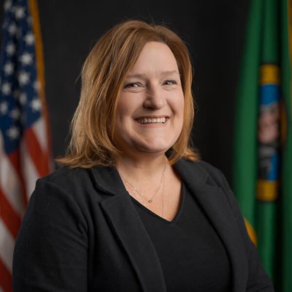 A woman with shoulder-length light brown hair, wearing a black blazer and top, smiles in front of the U.S. and Washington state flags.