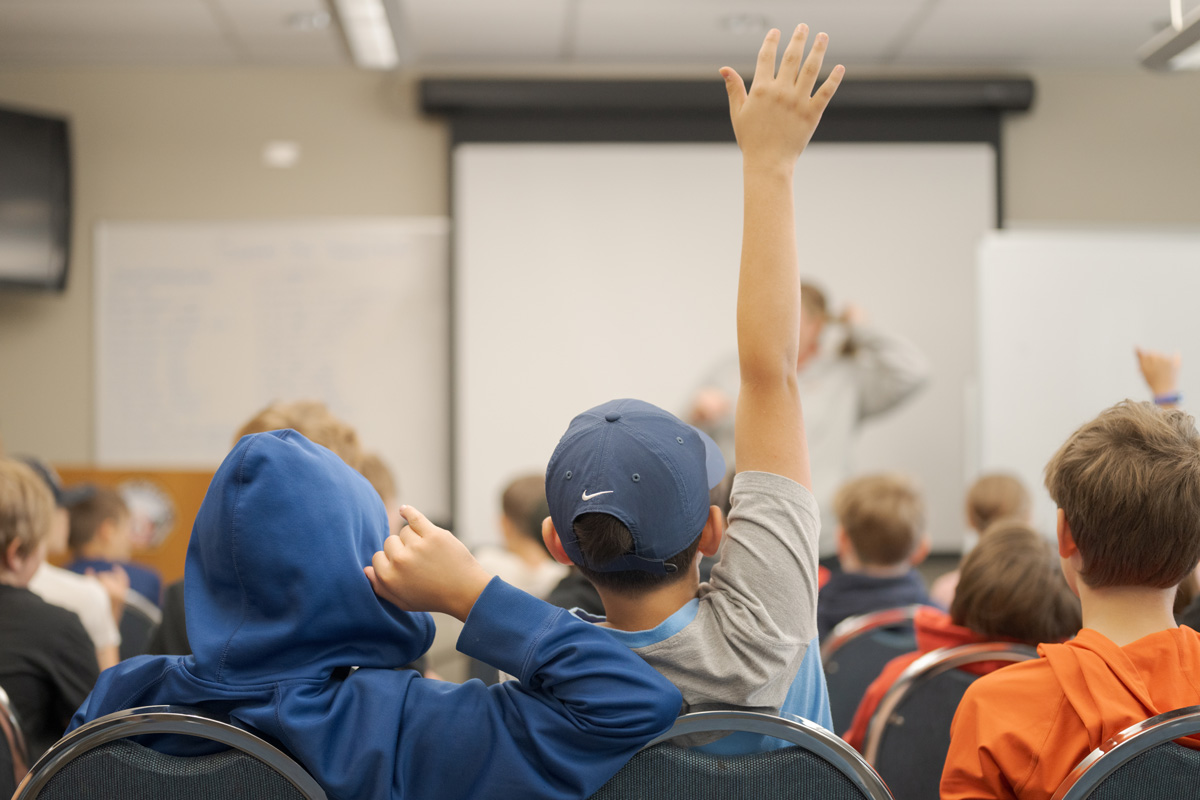 A group of students sit in a classroom facing a teacher, with one student raising their hand to ask or answer a question.