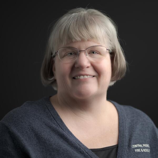 A middle-aged woman with short gray hair and glasses smiles at the camera, wearing a dark V-neck top with 