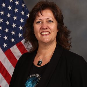 Woman smiling in front of an American flag, wearing a black blazer and a pendant necklace, with a neutral background.