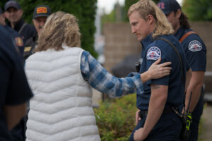 A woman in a white vest and plaid shirt touches the shoulder of a uniformed firefighter standing outside with other emergency personnel.