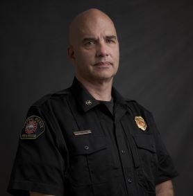 A man in a black uniform with badges stands against a dark background, facing the camera with a neutral expression.