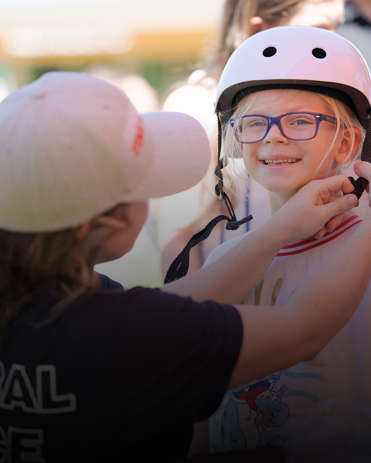 An adult fastens a white helmet on a smiling child wearing glasses and a T-shirt outdoors.