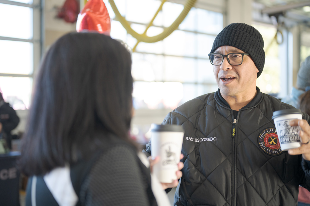 Two people stand indoors, holding coffee cups and talking. One wears a black beanie and a jacket with a Central Pierce Fire & Rescue budget and finance emblem, suggesting the setting is related to CPFR budget discussions at the fire station.
