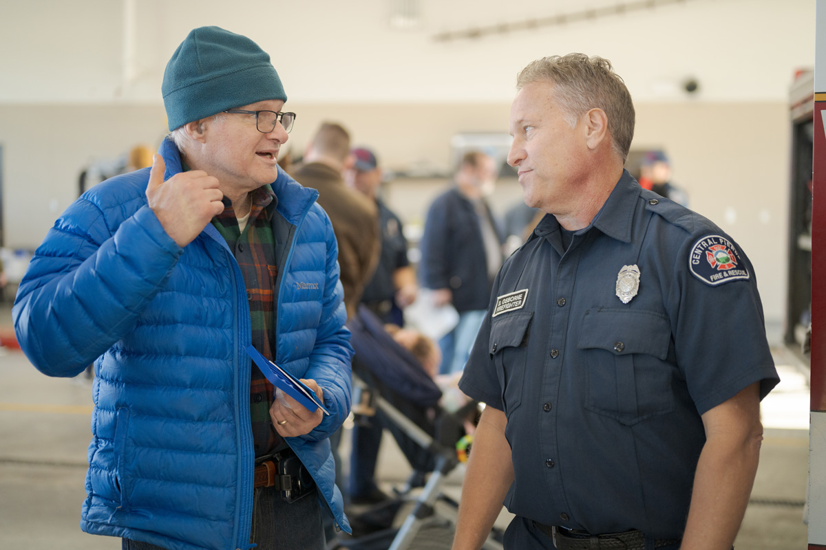 A man in a blue jacket and beanie talks to a uniformed fire department officer indoors, with other people and equipment visible in the background, possibly discussing the Central Pierce Fire & Rescue budget and finance.