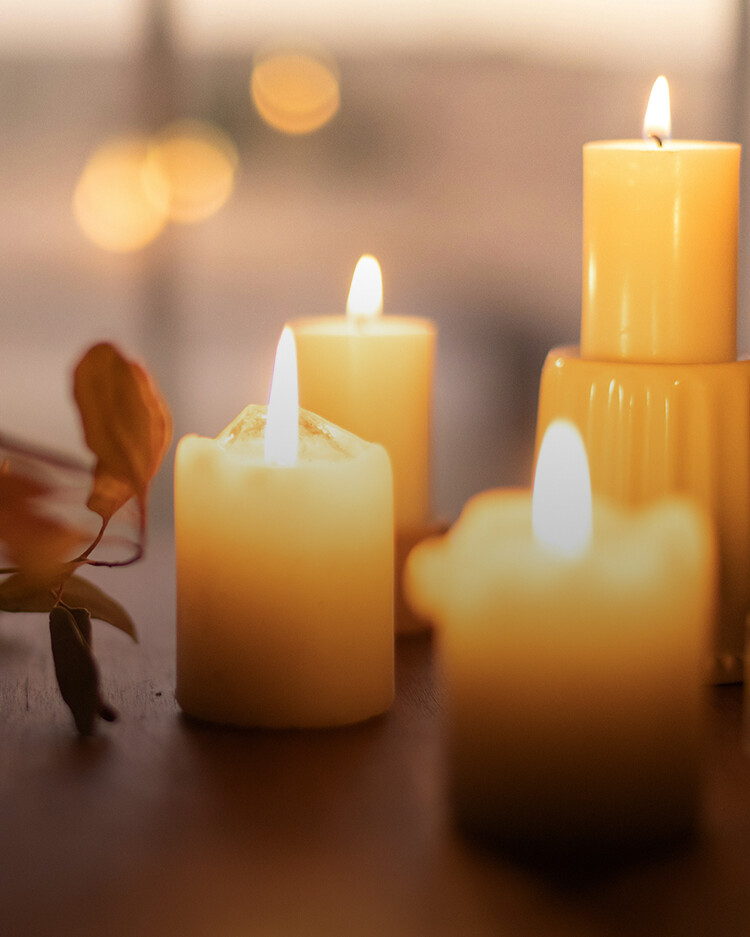 Four lit pillar candles on a wooden surface with blurred background and autumn leaves.