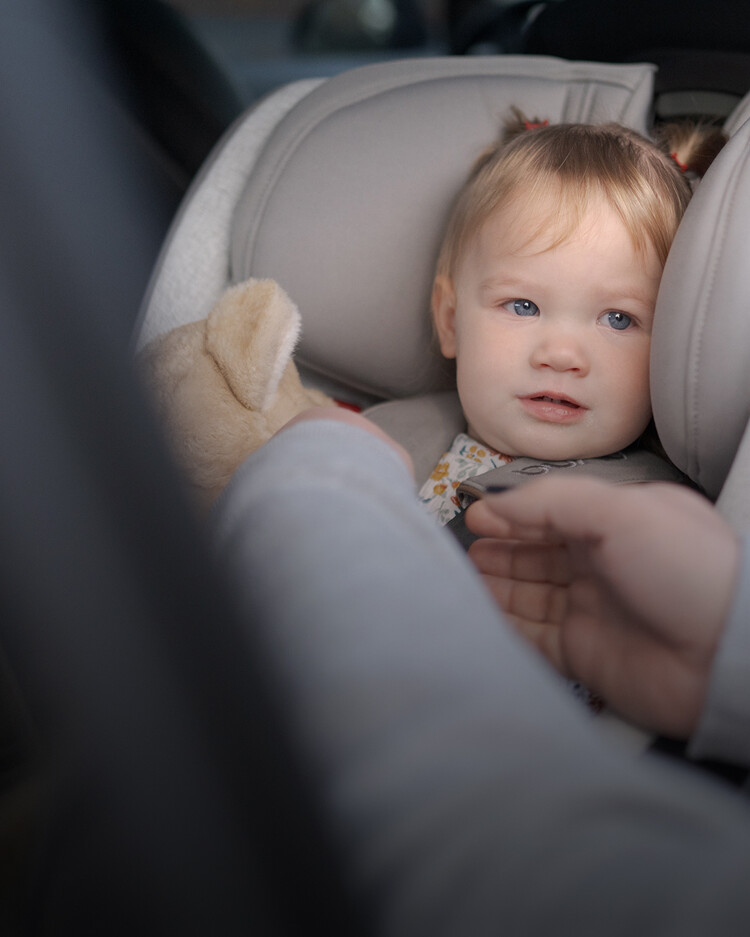 A toddler with light hair sits in a car seat, looking ahead, while an adult reaches toward them. A stuffed animal is partially visible beside the child.