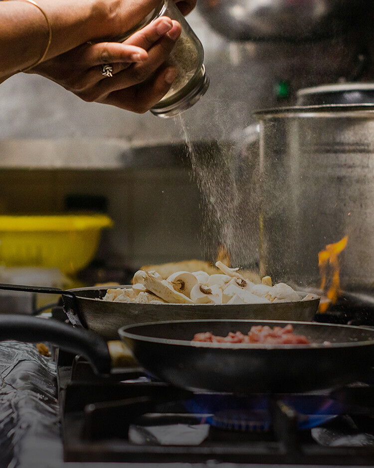 A hand is sprinkling seasoning onto sliced mushrooms in a frying pan on a gas stove, with another pan and kitchen utensils in the background.