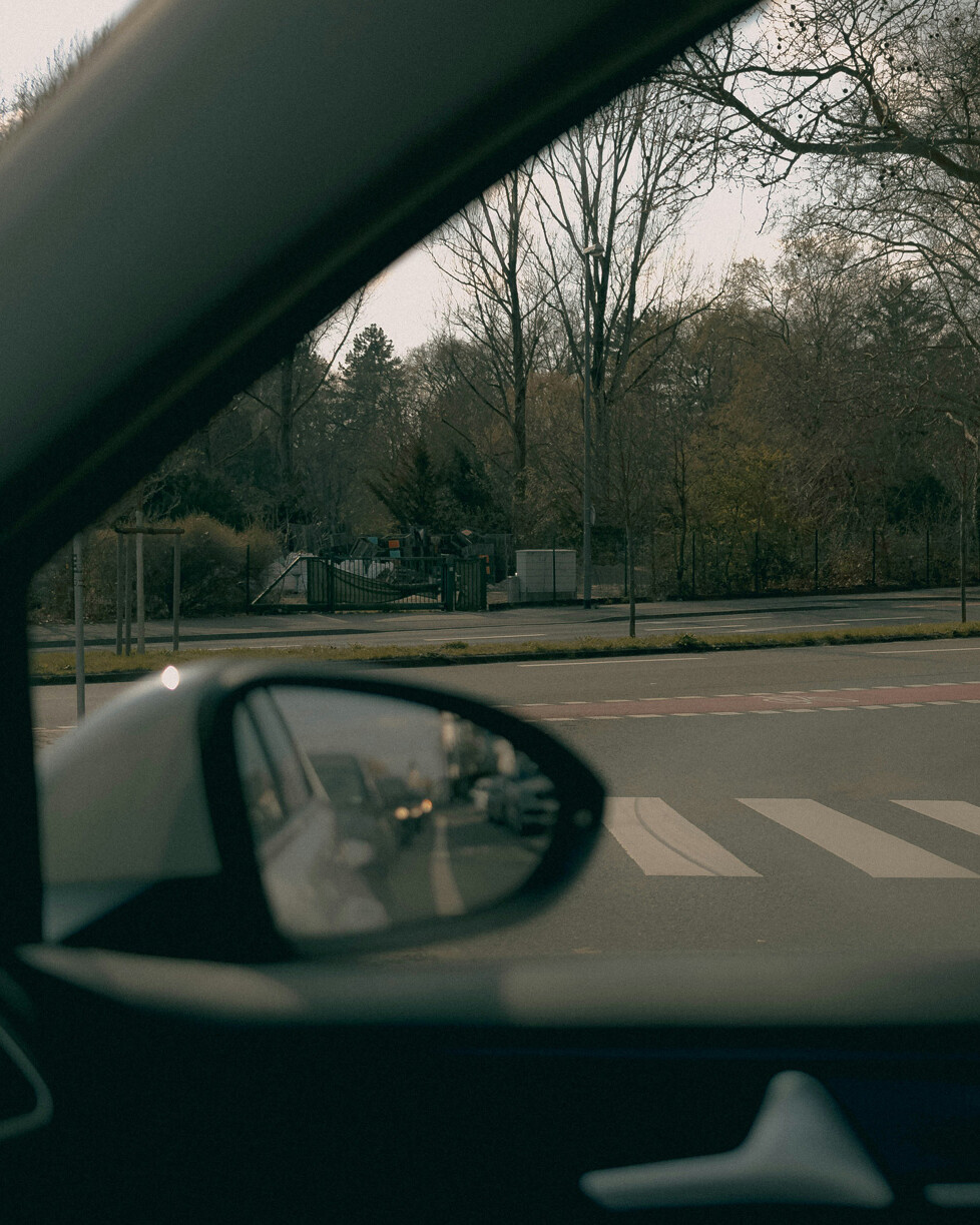 View from inside a car showing the side mirror, a road with a crosswalk, and trees in the background under an overcast sky.
