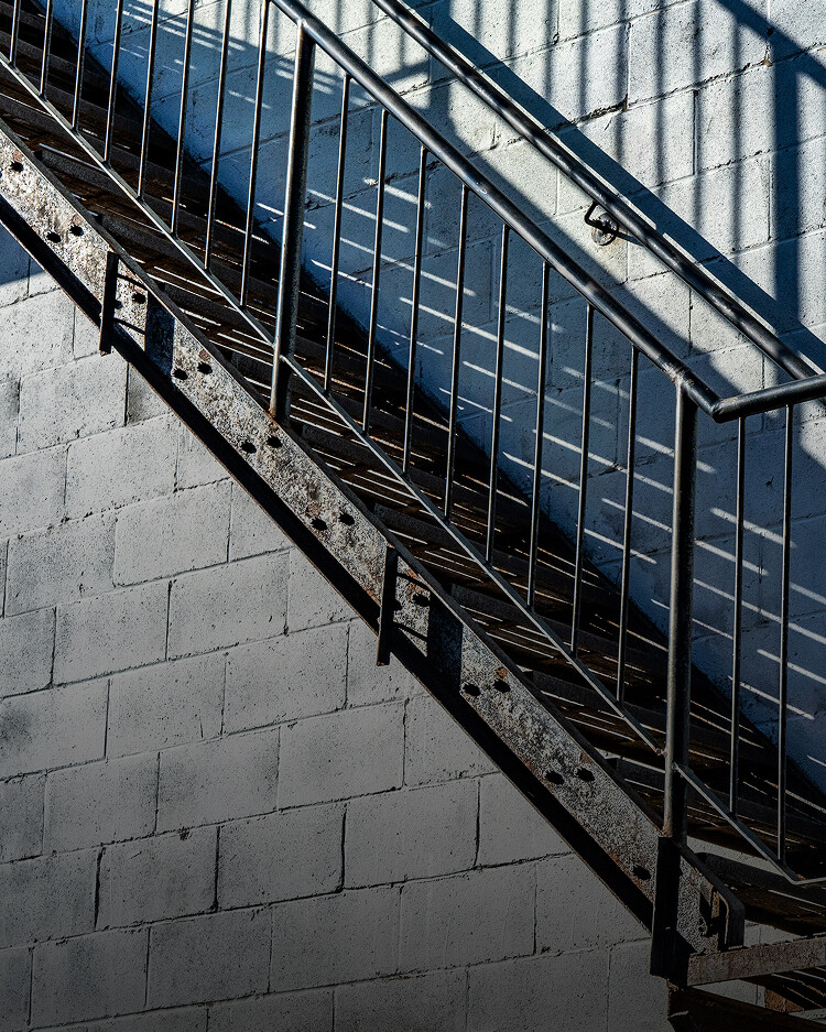 Metal staircase with railings angled upward against a white cinder block wall, casting geometric shadows from sunlight.