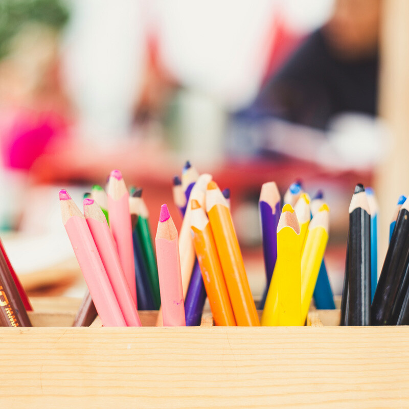 A wooden box holds a variety of colorful pencils, including pink, yellow, blue, black, and brown, arranged upright on a table.