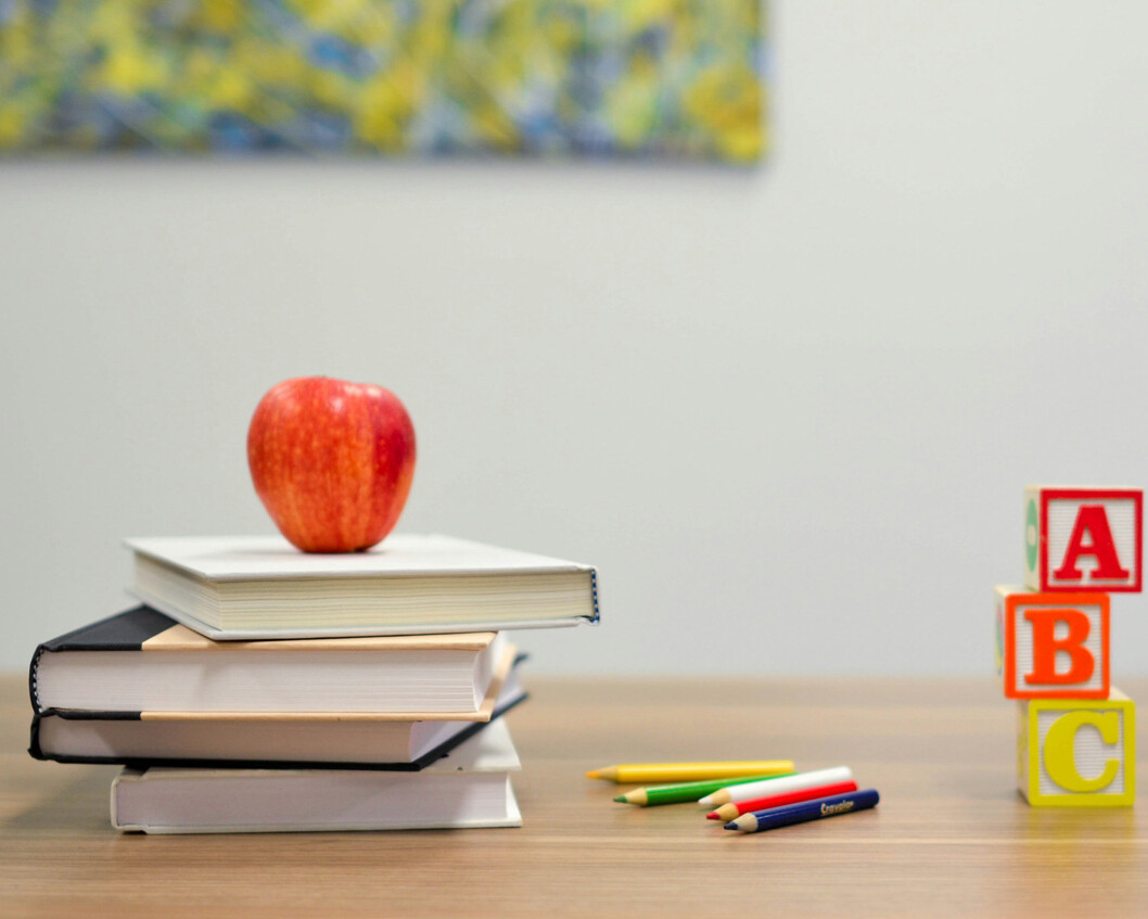 A red apple rests on a stack of three books beside colored pencils and three alphabet blocks spelling 