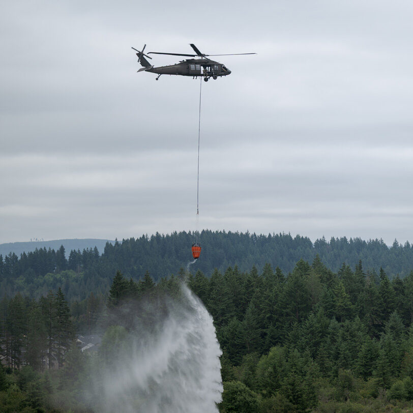 A Central Pierce Fire & Rescue helicopter drops water from a suspended bucket onto a forest fire below, with dense trees and cloudy skies in the background, highlighting wildfire awareness efforts.
