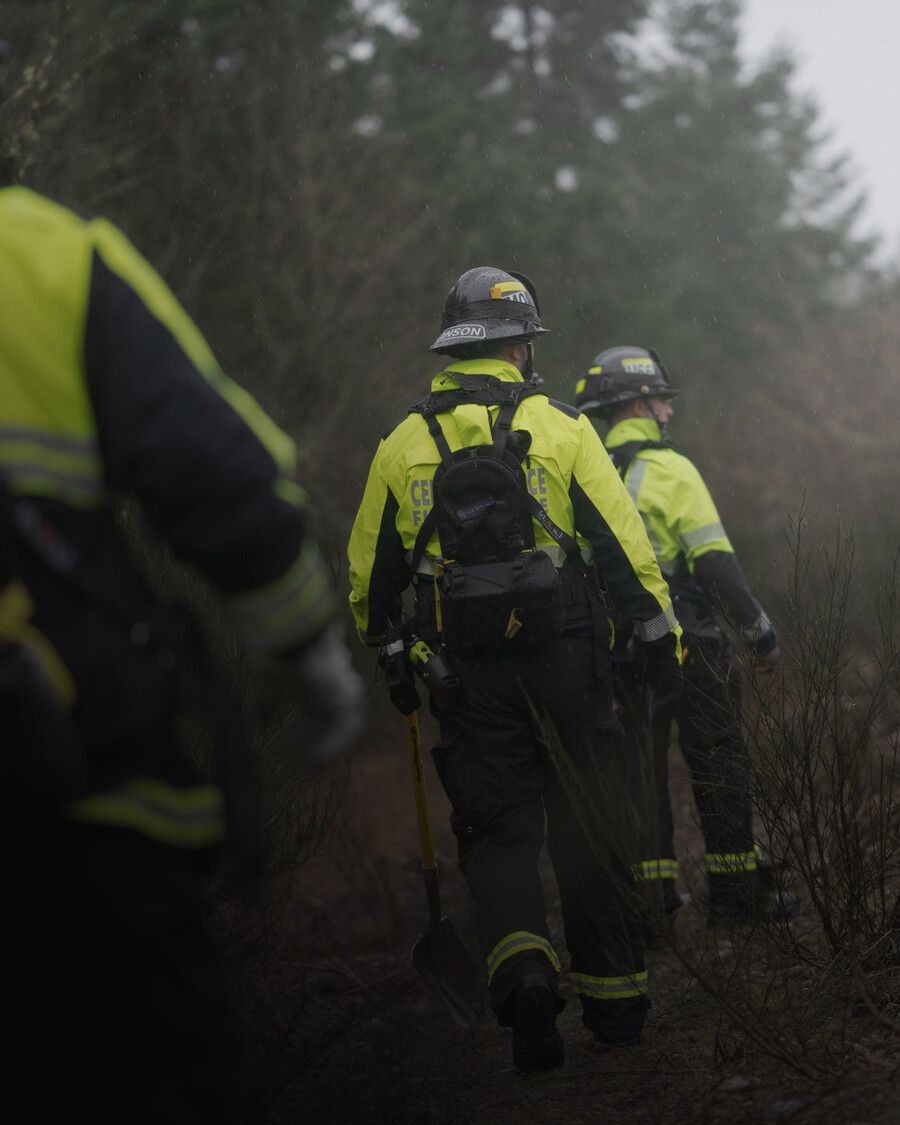 Three people in high-visibility jackets and helmets walk through a wooded area, with one carrying a shovel, highlighting Central Pierce Fire & Rescue wildfire awareness efforts.