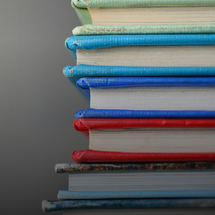 A close-up side view of a stack of hardcover books with colorful spines, arranged in a neat vertical pile against a plain background.