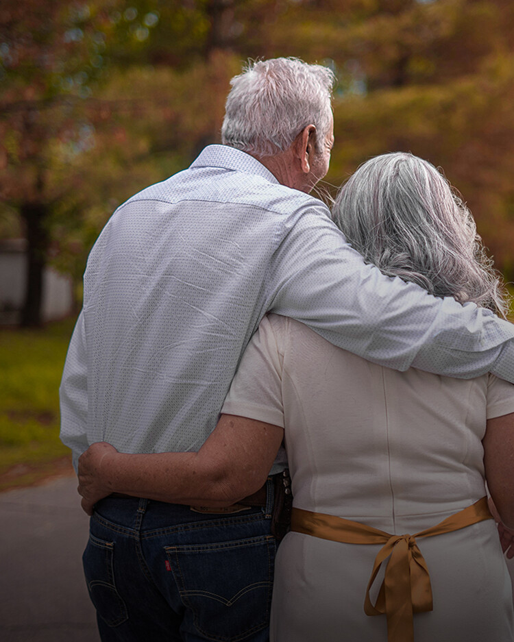 An elderly man and woman stand outdoors with their backs to the camera, the man’s arm around the woman’s shoulders as they walk together.