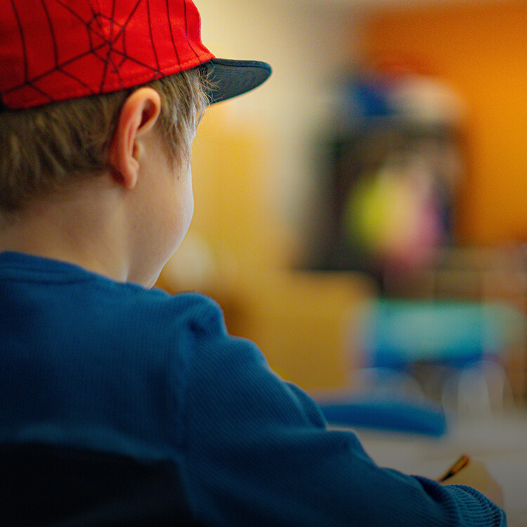 A child wearing a red cap sits at a desk, writing with a pencil, in a brightly lit classroom.