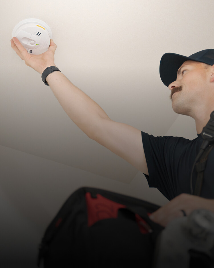 A person in a cap installs or checks a smoke detector on a ceiling while holding a bag.