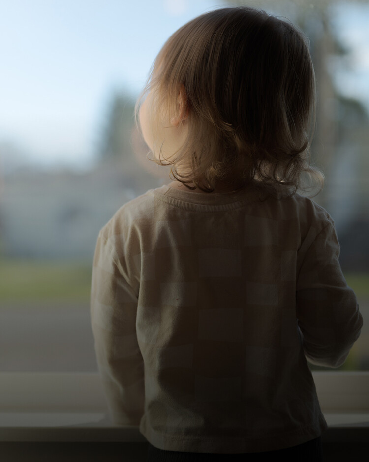 A young child with light brown hair stands indoors, looking out of a window with a blurred outdoor background.