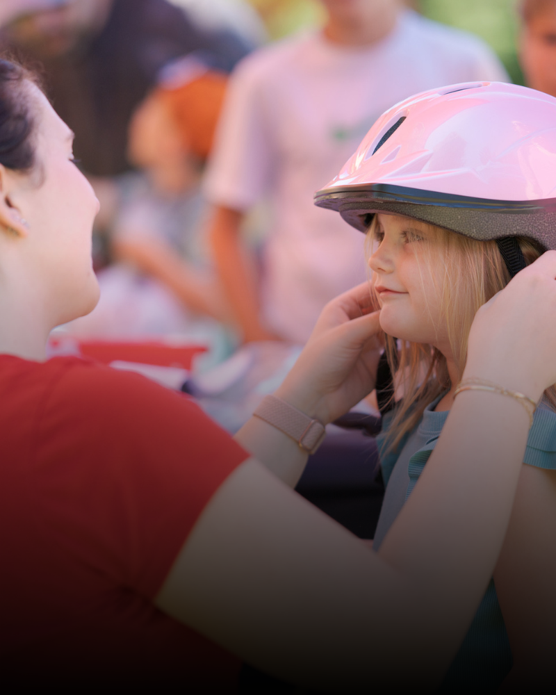 An adult helps a young girl put on a pink Central Pierce Fire & Rescue bike helmet while others stand in the background.