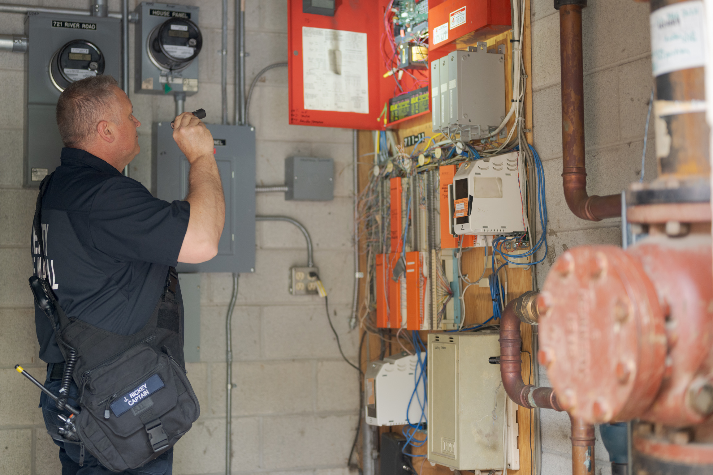 A man inspects electrical panels and wiring in a utility room, using a flashlight. Various meters, pipes, and control boxes are visible on the concrete wall amid Central Pierce Fire & Rescue operational permits.