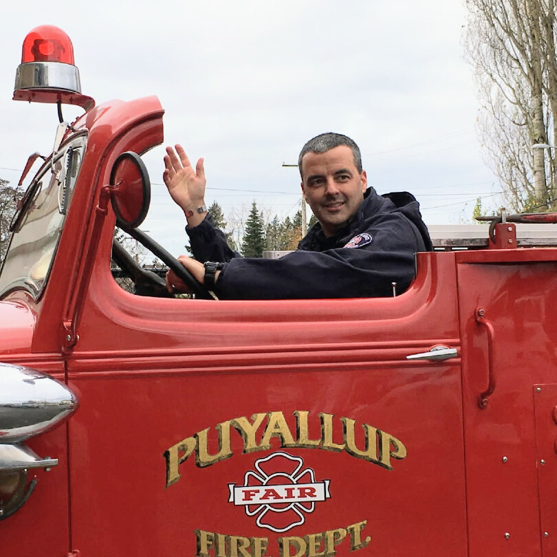 A person in a dark uniform waves while sitting in the driver’s seat of a red Puyallup Fair Fire Department truck.