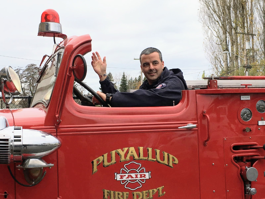 A person in a dark uniform waves while sitting in the driver&rsquo;s seat of a red Puyallup Fair Fire Department truck.