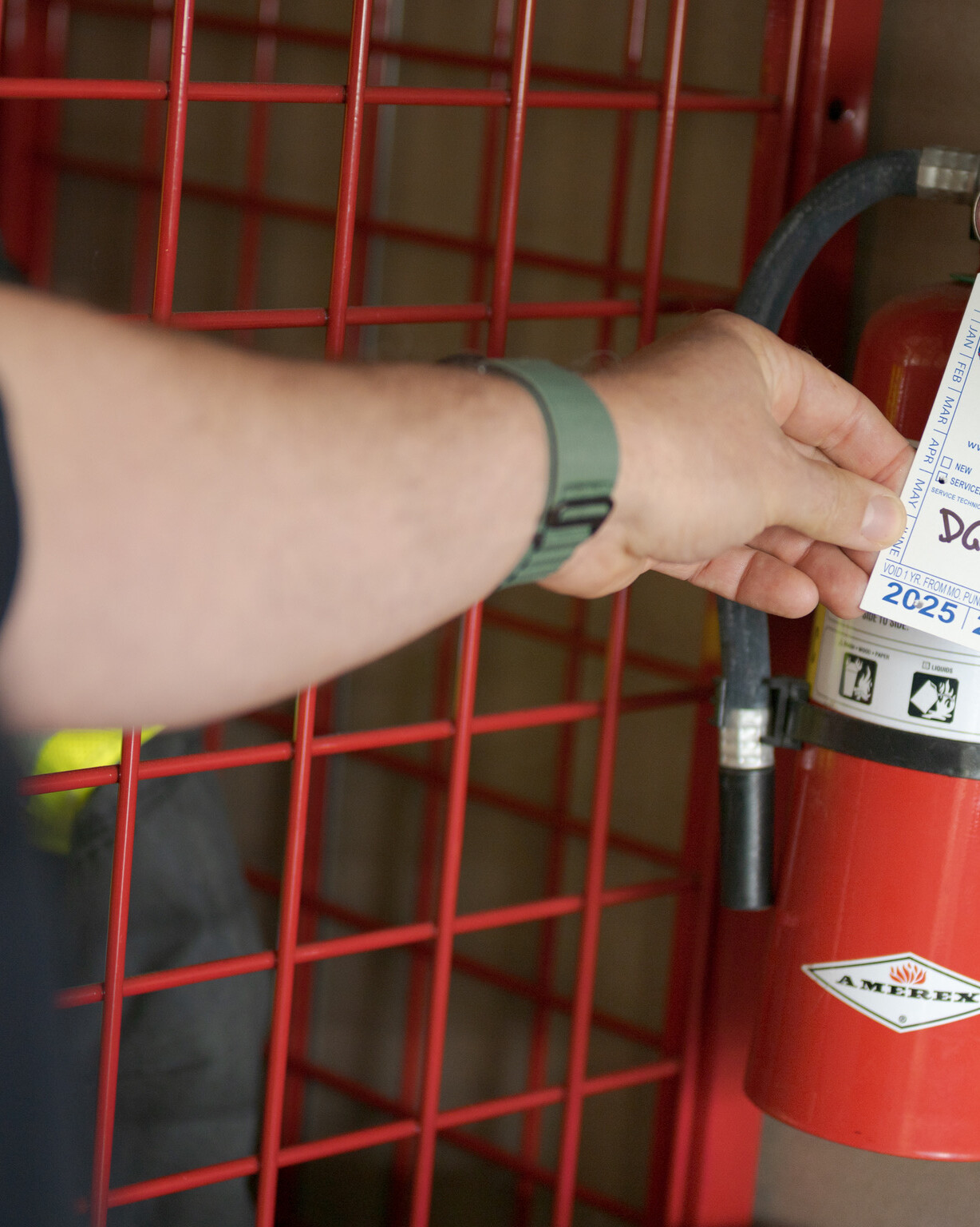 A person checks the inspection tag on a red Amerex fire extinguisher mounted on a wall with a red metal barrier, following guidance from Central Pierce Fire & Rescue fire extinguisher classes.