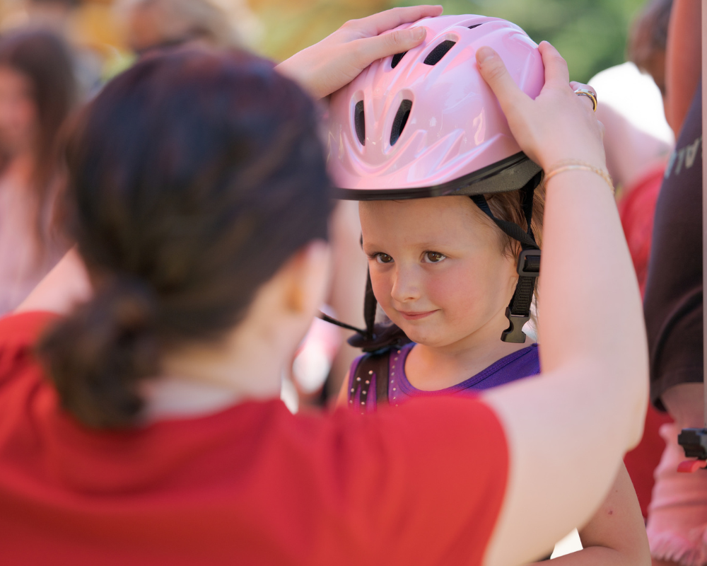 An adult helps a child put on a pink bicycle helmet outdoors, promoting CPFR driver and pedestrian safety as the child looks ahead while the helmet is adjusted.