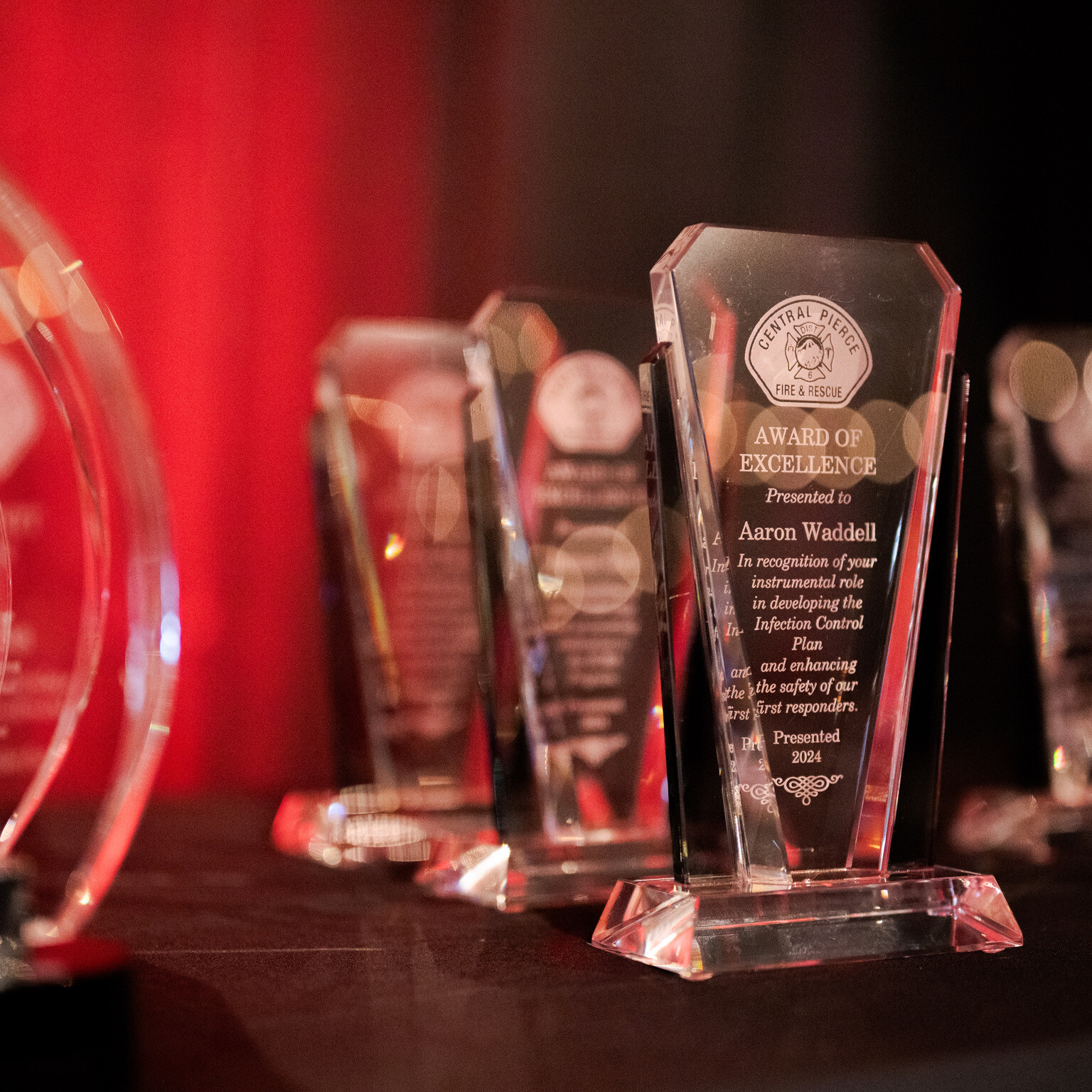 Several glass awards are displayed on a black tablecloth, with text and engravings visible on the plaques. A red and black background is out of focus.