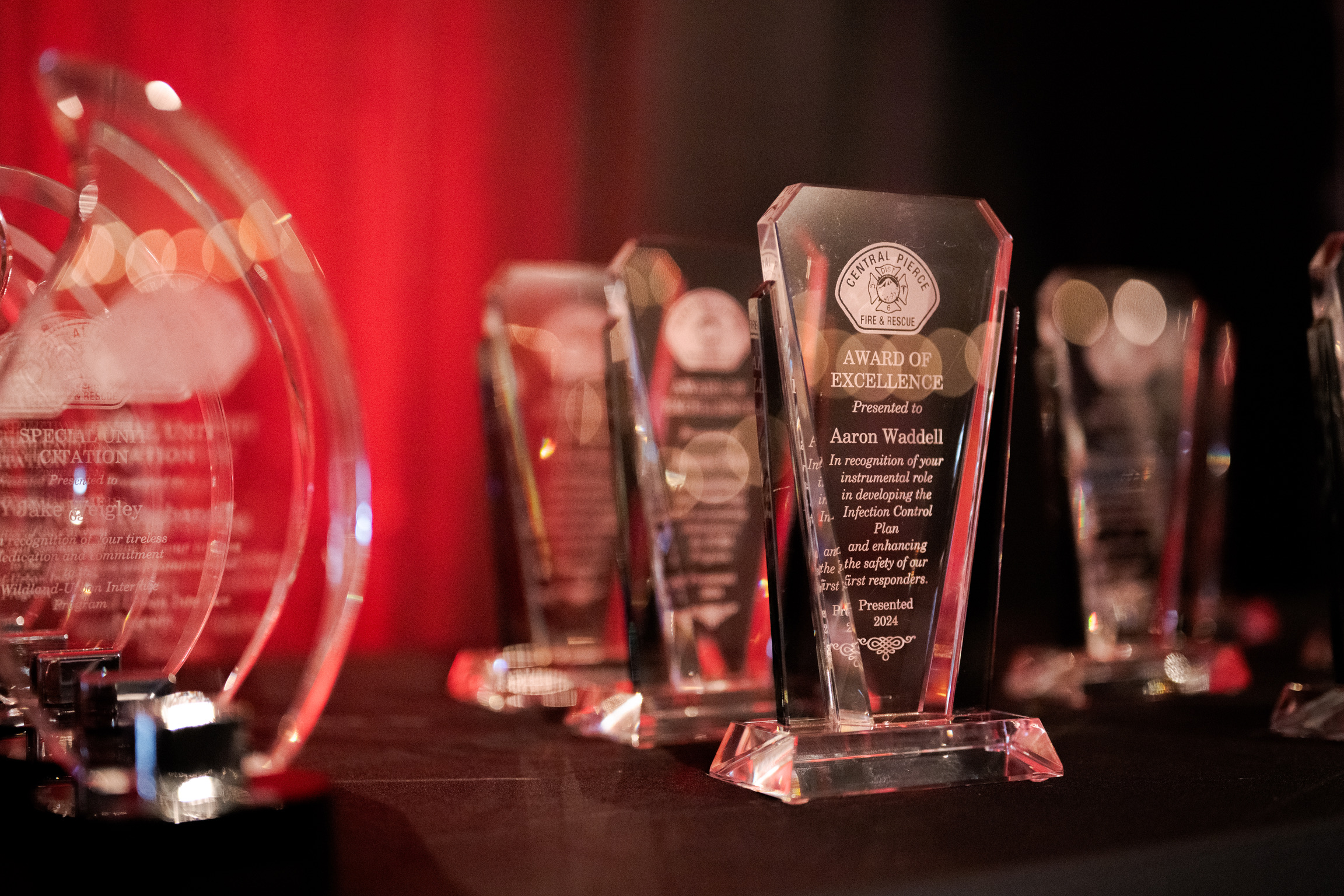 Several glass awards are displayed on a black tablecloth, with text and engravings visible on the plaques. A red and black background is out of focus.