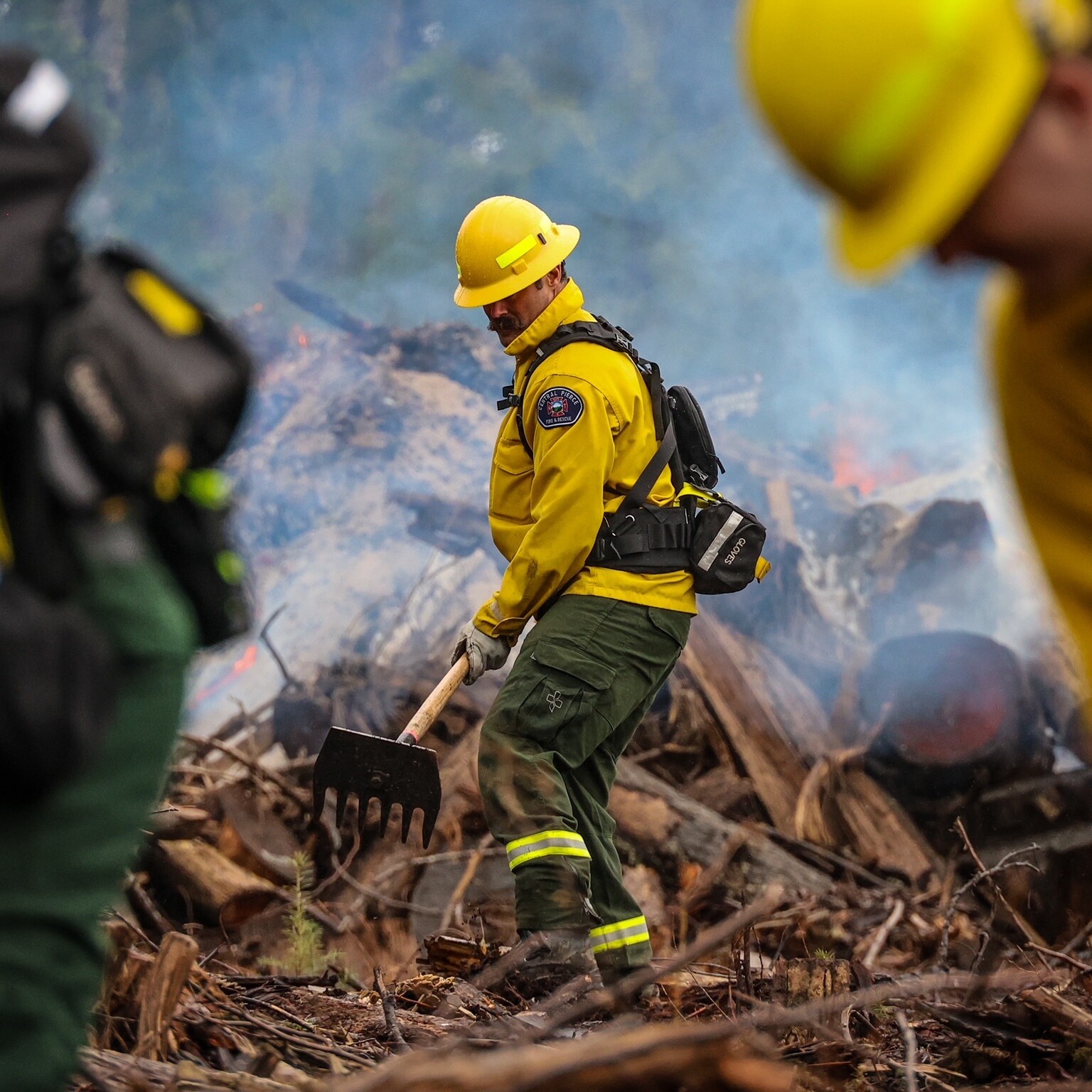 Firefighters in yellow protective gear work among debris and smoke, with one using a firefighting tool near a smoldering area in a forested environment.