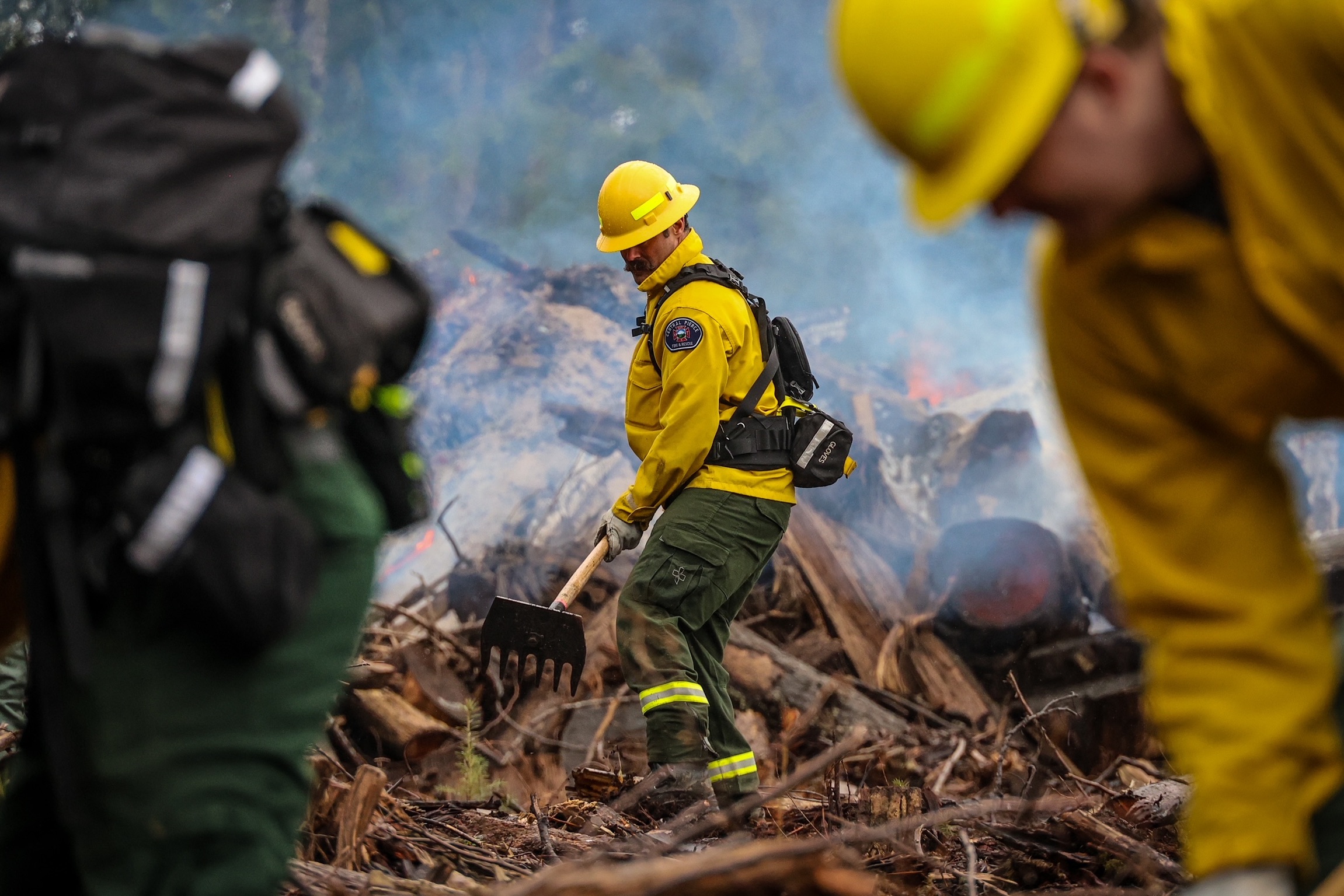 Firefighters in yellow protective gear work among debris and smoke, with one using a firefighting tool near a smoldering area in a forested environment.