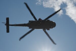 A black helicopter seen from below flies against a blue sky, with sunlight and some clouds in the background.