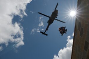 A helicopter hovers in the sky while two people are lowered on a cable near a building, with bright sunlight and clouds in the background.