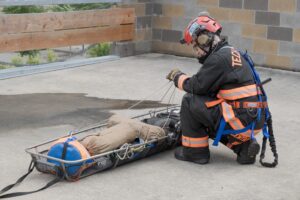 A firefighter in safety gear kneels beside a rescue stretcher with a training dummy secured inside, preparing ropes for a rescue exercise on a concrete surface.