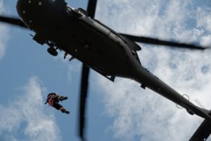 A person in an orange suit is being hoisted up by a cable to a black helicopter flying overhead against a partly cloudy sky.