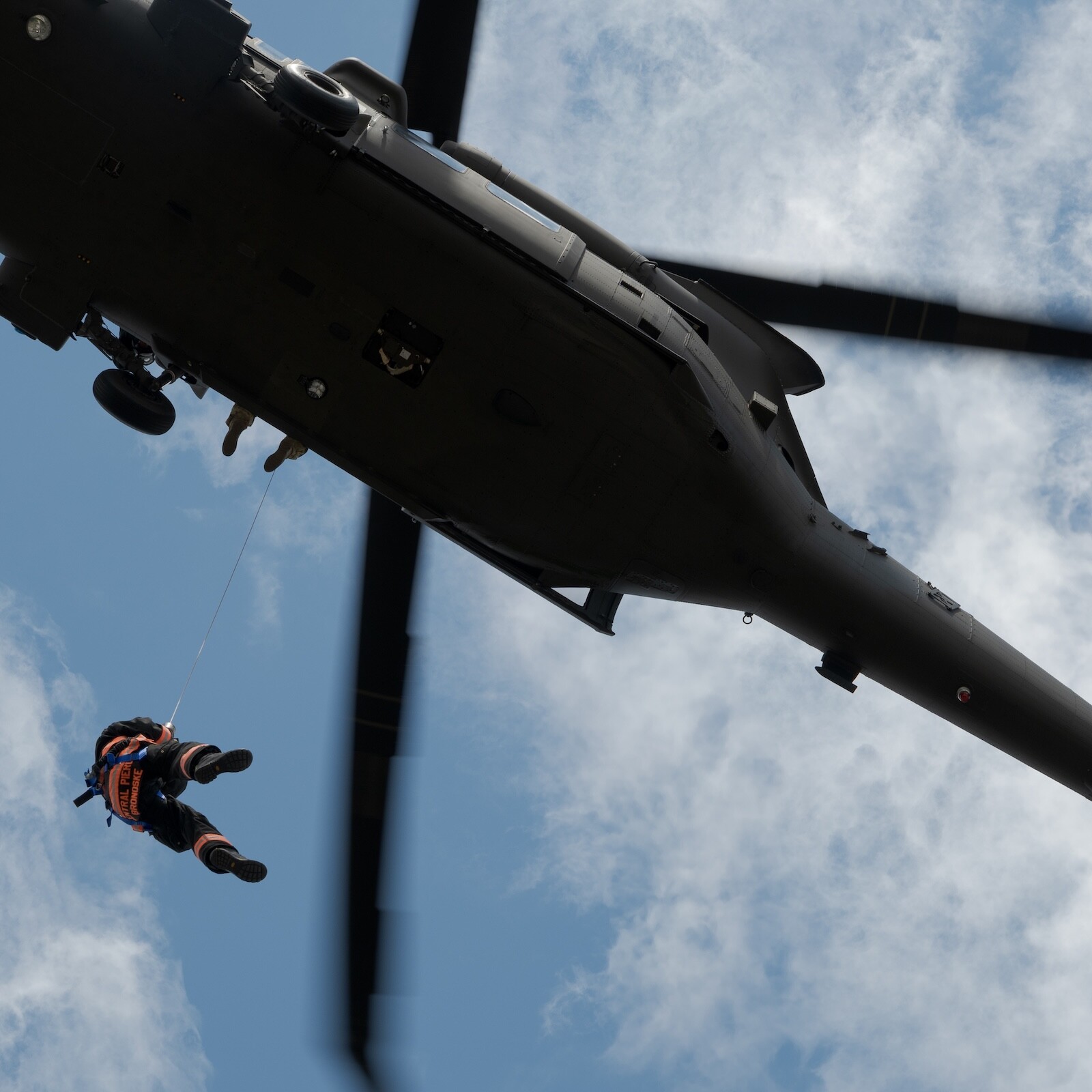 A person in an orange suit is being hoisted up by a cable to a black helicopter flying overhead against a partly cloudy sky.