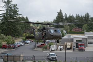 A military helicopter hovers above a parking lot near a building, with emergency vehicles and trees in the background.