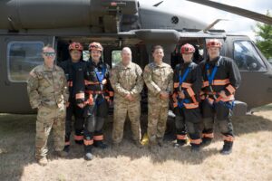 Six people in uniforms, three in camouflage and three in black with orange safety gear, stand in front of a military helicopter on grass.