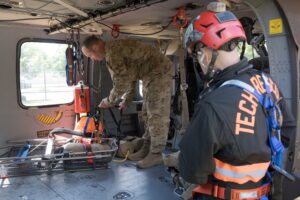 A soldier and a rescue technician prepare equipment inside a helicopter, with rescue gear and harnesses visible.