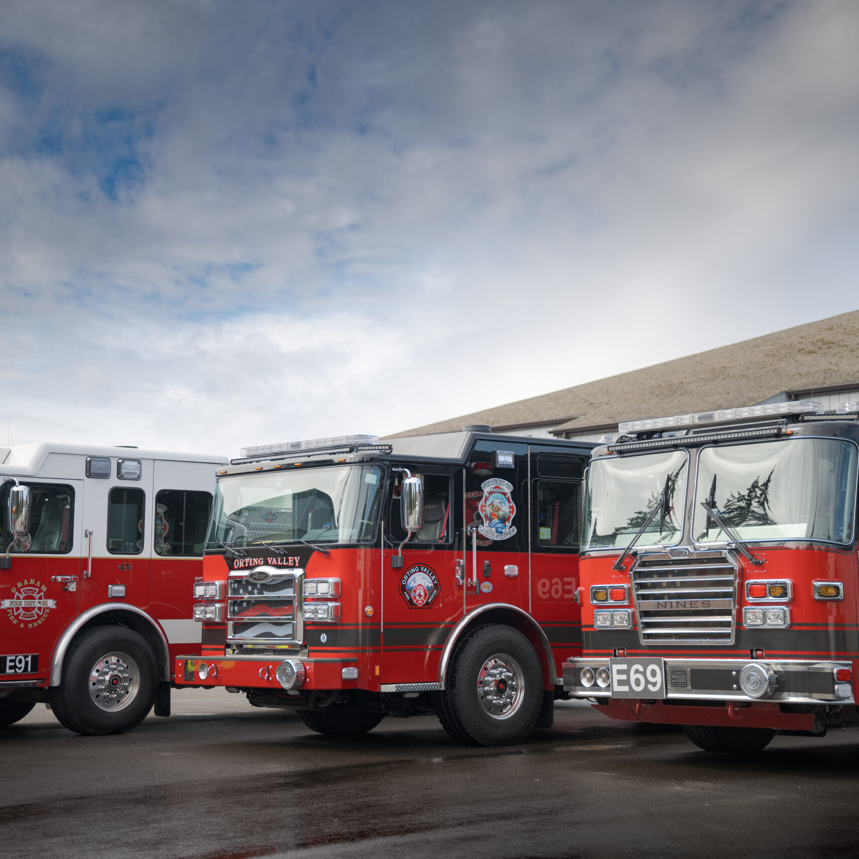 Three red fire trucks are parked side by side outside a building under a partly cloudy sky.