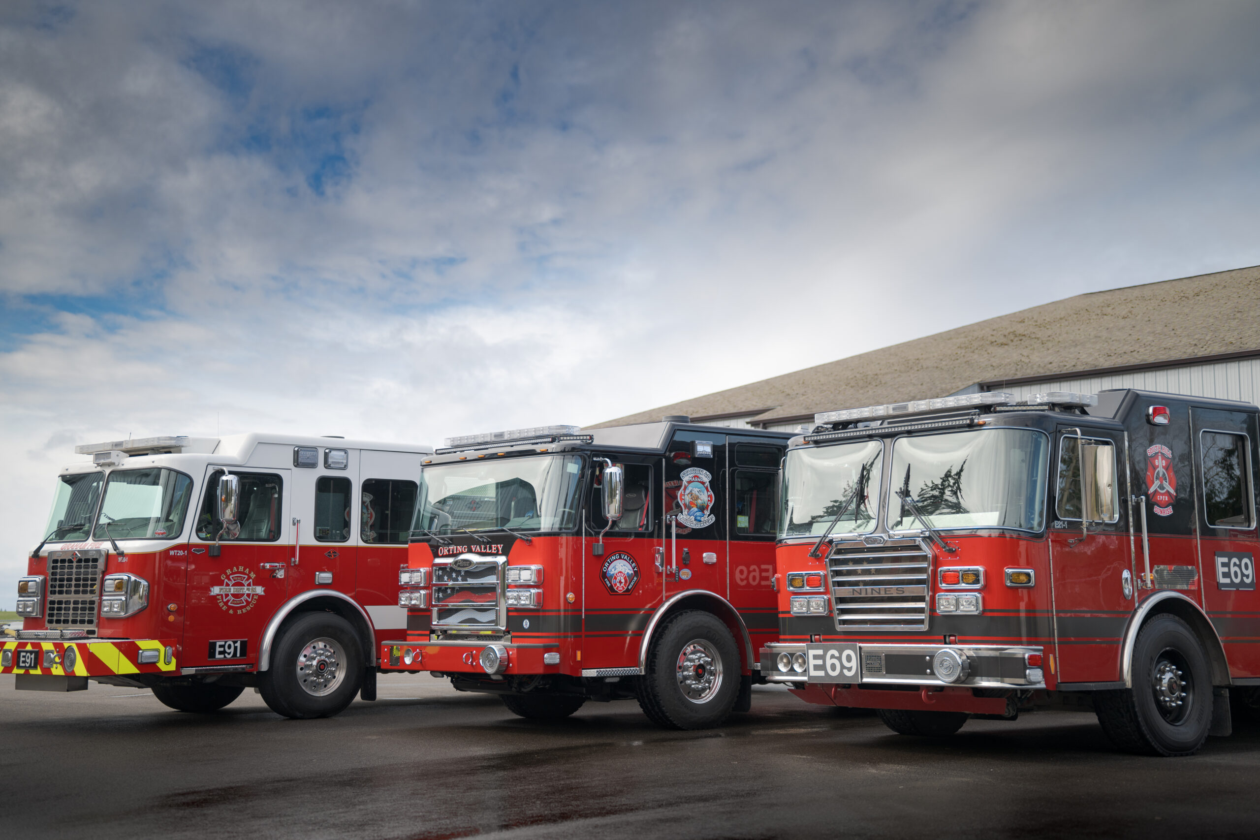 Three red fire trucks are parked side by side outside a building under a partly cloudy sky.