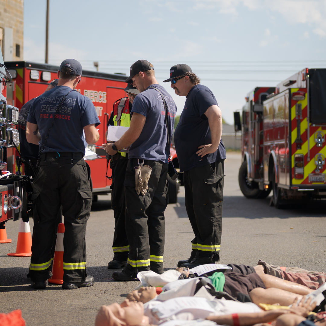 Four firefighters stand beside fire trucks and orange cones in a parking lot, with mannequins on the ground in the foreground, suggesting a training exercise or drill.