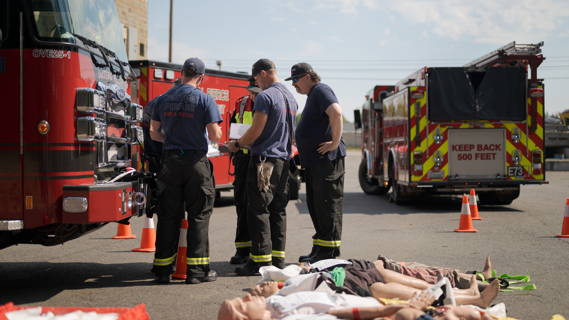 Four firefighters stand beside fire trucks and orange cones in a parking lot, with mannequins on the ground in the foreground, suggesting a training exercise or drill.